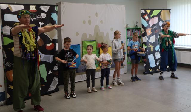 Na zdjęciu znajduje się kobieta i mężczyzna przebrani za smieciaki i mają ręce wyciągnięte przed siebie, pomiędzy nimi stoi piątka dzieci, którzy grają na instrumentach. W tle znajdują się trzy tablice przedstawiające śmieci oraz odpady.