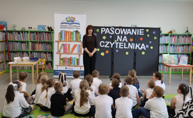 Na zdjęciu znajdują się przedszkolaki siedzące na dywanie, przed nimi stoi kobieta. W tle znajduje się czarna tablica z napisem, baner z książkami i logiem biblioteki, stolik i półki z książkami.