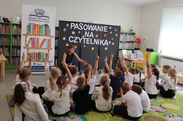 Na zdjęciu znajdują się dzieci siedzące na dywanie, podnoszą ręce do góry, przed nimi klęka kobieta i wskazuje ręką, w tle znajduje się czarna tablica z napisem, baner z logiem biblioteki i książkami oraz półki z książkami.