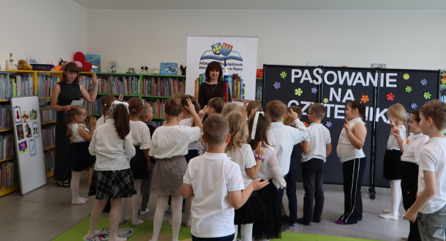 Na zdjęciu znajdują się stojące dzięci, mają podniesione ręce do góry, przed nimi stoją dwie kobiety, a w tle znajduje się czarna tablica z napisem, baner z logiem biblioteki i książkami, półki z książkami.