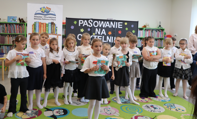 Na zdjęciu znajduje się grupa dzieci, które stoją obok siebie na dywanie, przed nimi stoi dziewczynka i mówi wiersz. Dzieci trzymają w ręku książki oraz dyplomy. W tle znajdują się półki z książkami, baner z logiem biblioteki i kksiążkami oraz czarna tablica z napisem.