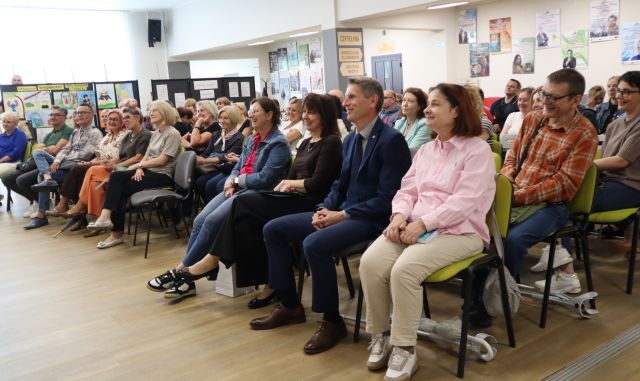 Na zdjęciu znajduje się hol biblioteczny ze stuosobową widownia. W tle znajdują się okna, plakaty na ścianach.