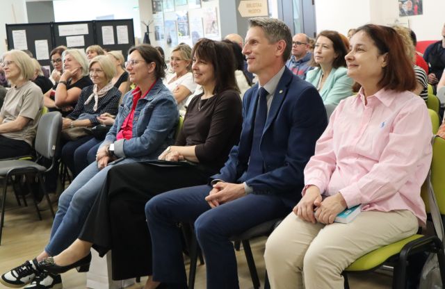 Na zdjęciu w centrum znajduje się pierwszy rząd widowni składający się z czterech osób, a w tle znajdują się inne osoby siedzące na widowni na holu biblioteki.