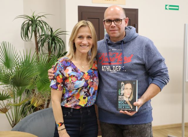 Na zdjęciu znajduje się kobieta z mężczyzną, który ma książkę w ręce , w tle znajdują się drzwi oraz palma.