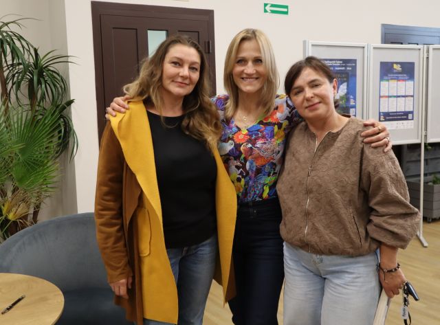 Na zdjęciu znajdują się trzy kobiet stojące wspólnie do zdjęcia. W tle znajdują się drzwi oraz palmy oraz plakaty.
