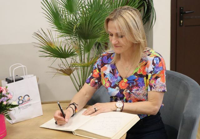 Na zdjęciu znajduje się kobieta siedząca przy stoliku i wpisująca się do księgi, na stoliku leżą kwiaty i upominek. W tle znajdują się drzwi oraz palmy.