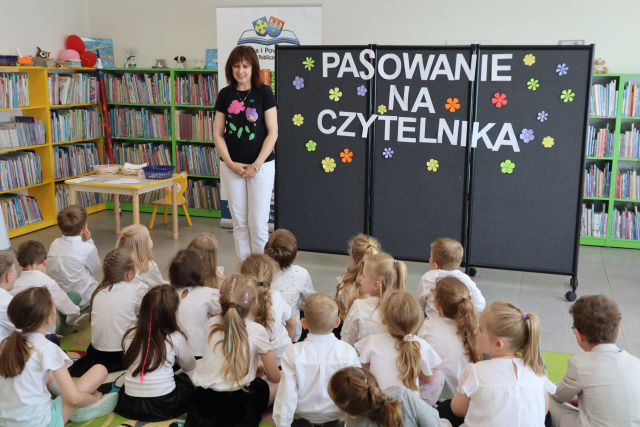 Na zdjęciu znajdują się siedzące na dywanie przedszkolaki, przed nimi stoi kobieta, za nią znajduje się stolik, baner z logiem biblioteki, po prawej stronie czarna tablica z napisem pasowanie na czytelnika. W tle znajdują się półki z książkami.