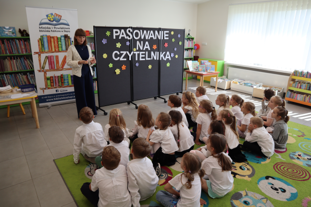 Na zdjęciu znajdują się dzieci siedzące na dywanie, przed nimi stoi kobieta, za nią stoi baner z logiem biblioteki i książkami, po lewej stronie stolik, po prawej stronie tablica z napisem. W tle znajdują się półki z książkami oraz okna.