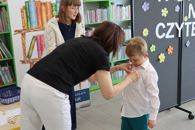 Na zdjęciu znajdują się dwie kobiety, jedna z nich się nachyla i nakleja chłopcu kartkę z imieniem. W tle znajduje się baner z logiem biblioteki i książkami oraz czarna tablica z napisem.