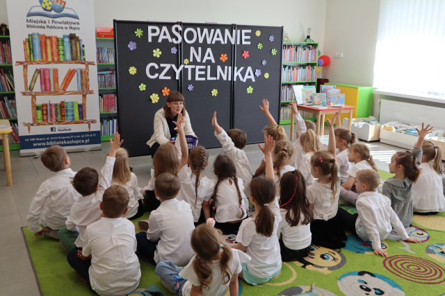 Na zdjęciu znajdują się dzieci na dywanie, część z nich ma podniesioną rękę do góry, prze nimi kuca kobieta z koszyczkiem w ręce. Za kobietą znajduje się baner z logiem biblioteki i książkami, czarna tablica z napisem, a w tle półki z książkami i okna po prawej stronie.