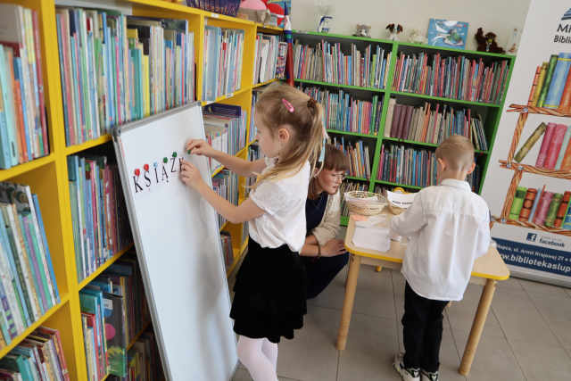 Na zdjęciu po lewej stronie dziewczynka przypina do białej tablicy literkę, obok kuca kobieta i pomaga chłopcu przy stoliku z ułożeniem wyrazu. W tle znajdują się półki z książkami oraz po prawej stronie baner z książkami i logiem biblioteki.