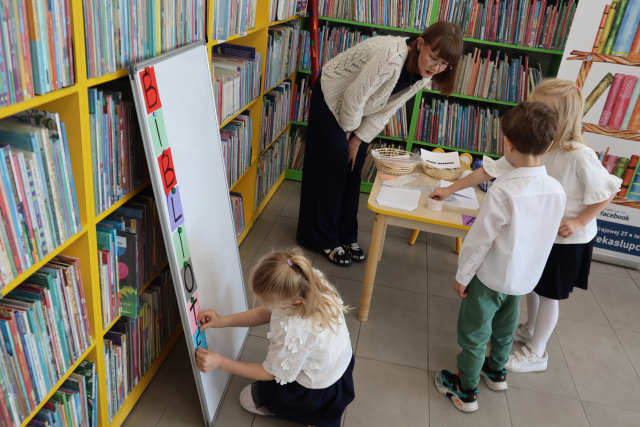 Na zdjęciu po lewej stronie dziewczynka przypina kolorową literkę do białej tablicy, która jest oparta o półki z książkami, po prawej stronie przy stoliku stoi dwójka dzieci oraz nachyla się kobieta. W tle znajduje sie baner z książkami oraz półki z książkami.