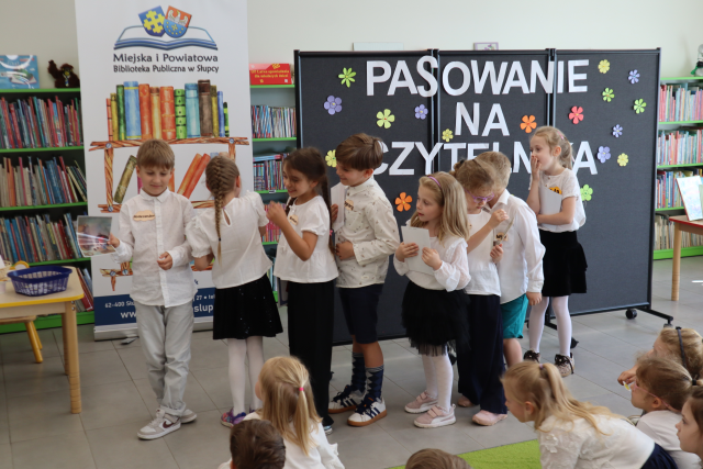 Na zdjęciu znajduje się siódemka dzieci, które mają w ręku rysunki, przed nimi siedzi grupa na dywanie. W tle znajduje się czarna tablica z napisem, baner z logiem biblioteki i książkami, półki z książkami oraz stolik po lewej stronie.