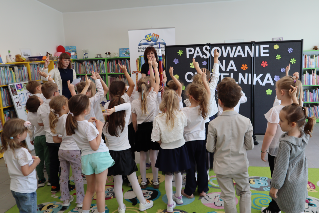 Na zdjęciu znajduje się grupa przedszkolaków stojąca na dywanie z podniesioną prawą ręką, przed nimi stoją dwie kobiety, w tle znajduje się czarna tablica z napisem, baner z logiem biblioteki oraz półki z książkami.