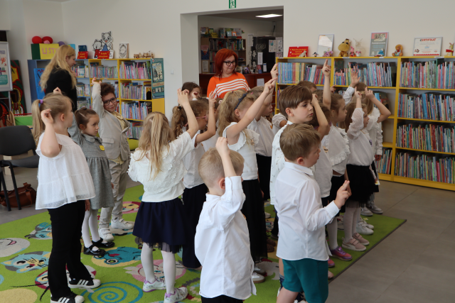 Na zdjęciu znajdują się dzieci stojące na dywanie z podniesioną do góry ręką, za nimi stoją dwie kobiety. W tle znajdują się półki z książkami.