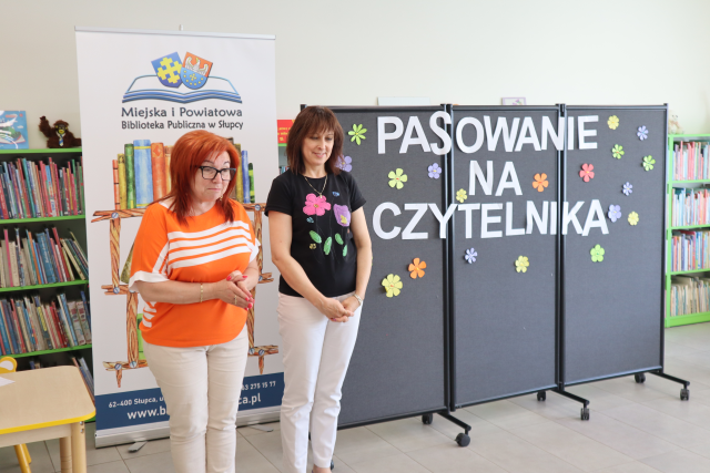 Na zdjęciu znajdują się dwie kobiety stojące na tle banera z książkami oraz czarnej tablicy z napisem pasowanie na czytelnika, w tle znajdują się półki z książkami.