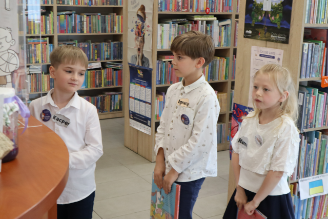 Na zdjęciu znajduje się trójka dzieci stojąca w kolejce do biurka aby wypożyczyć trzymaną przez siebie książkę. W tle znajdują się półki z ksiażkami.