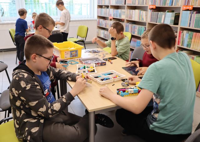 Na zdjęciu znajduje się ośmiu chłopców, piątka układa klocki LEGO przy dwóch złączonych stolikach, a trójka przy jednym obok. W tle znajdują się półki z książkami.