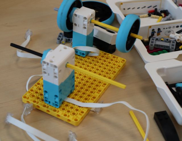 Na zdjęciu znajduje się konstrukcja z LEGO posiadająca kółka i leży na stoliku, obok plastikowego pudełka z klockami.