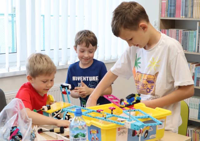 Na zdjęciu znajduje się trójka chłopców i układają klocki LEGO przy jednym stoliku, na którym leżą plastikowe pojemniki z klockami LEGO. W tle znajdują sie okna i półki z książkami.