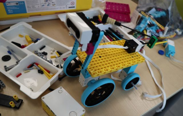Na zdjęciu znajduje się stojący na stoliku robotyczny pies z klocków LEGO z kółkami, dookoła niego znajdują się klocki LEGO w plastikowych pojemnikach.