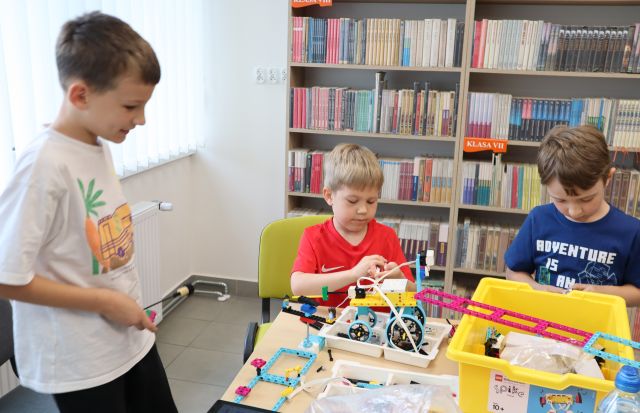 Na zdjęciu znajduje się trójka chłopców przy stoliku, którzy wspólnie doskonalają swoją konstrukcje z klocków LEGO. W tle znajdują się półki z książkami i okna po lewej stronie.