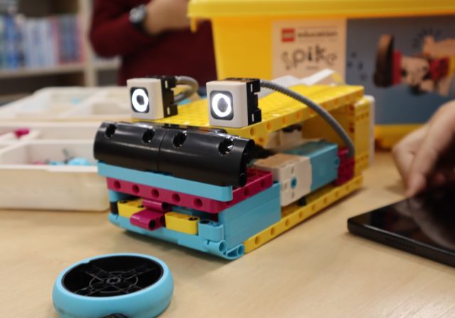 Na zdjęciu znajduje się stojąca na stole zbudowana z klocków LEGO robotyczna żaba. W tle na stoliku znajdują się klocki LEGO w plastikowych pojemnikach.