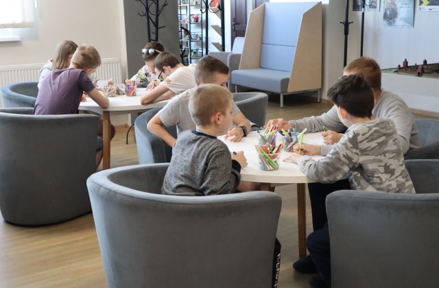Na zdjęciu znajduje się młodzież przy dwóch okrągłych stolikach na holu biblioteki i rozwiązują zadania. W tle znajduje się makieta miasta oraz kanapa.