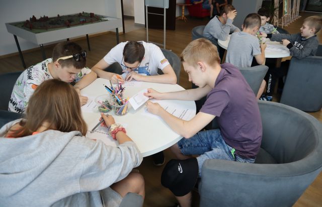 Na zdjęciu znajduje się czwórka młodzieży, którzy rozwiązują zakodowane wiadomości przy okrągłym stole, na holu biblioteki. W tle znajduje się drugi stolik, przy którym siedzi młodzież oraz makieta miasta.