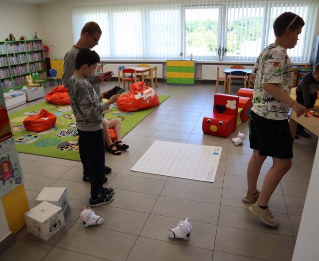 Na zdjęciu znajduje się trójka chłopców, którzy przy pomocy tabletów sterują robotami, na podłodze leży mata. W tle znajduje się dywan, pufy, pociąg z puf, małe stoliczki oraz okna.