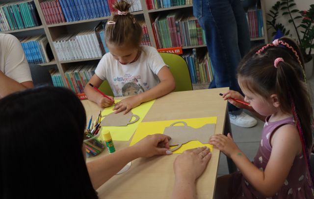 Na zdjęciu znajdują się dwie dziewczynki przy stole, które odrysowują na żółtej kartce puchar, dziewczynce po prawej pomaga mama. W tle znajdują się półki z książkami.