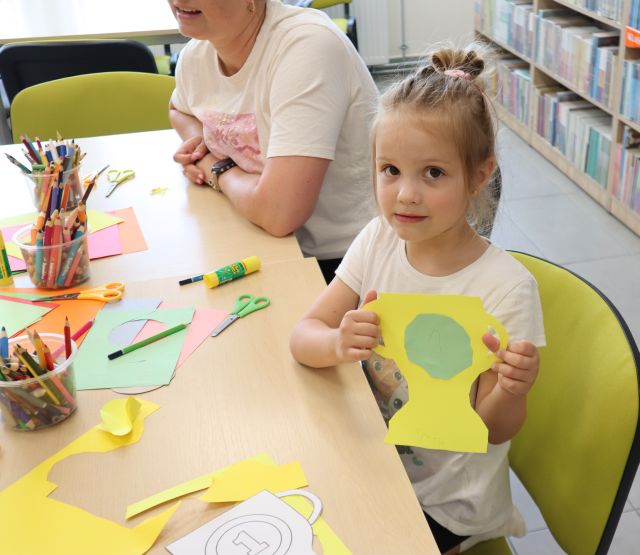 Na zdjęciu znajduje się dziewczynka, która siedzi przy stoliku i trzyma w ręce przed sobą zrobiony z papieru puchar dla taty, za nią siedzi kobieta. Na stoliku znajdują się kartki, kredki, nożyczki. W tle znajduje się fragment półki z książkami.