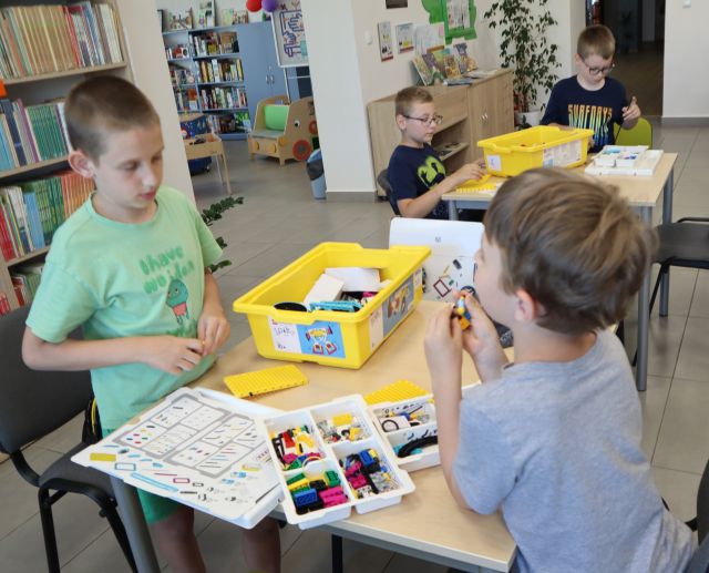 Na zdjęciu znajduje się dwóch chłopców przy stoliku i układają znajdujące się na nim klocki LEGO, w tle przy osobny stoliku siedzi dwóch chłopców, którzy również układają klocki przy stoliku. W tle znajdują się półki z książkami, szafki.