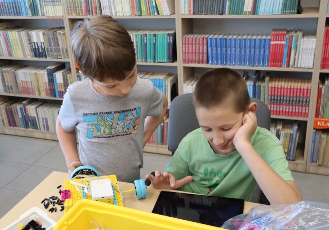 Na zdjęciu znajduje się dwóch chłopców jeden siedzi, drugi stoi obok niego i razem programują na tablecie swojego robota. Na stoliku przed nimi znajdują się klocki, a w tle półki z książkami.