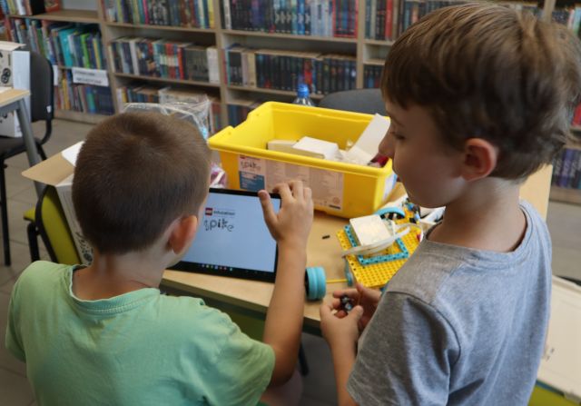Na zdjęciu znajduje się dwóch chłopców przy stoliku, mają przed sobą tableta na którym kodują swoją konstrukcję z klocków LEGO, na stoliku leżą pudełka z klockami, a w tle znajdują się półki z książkami. Zdjęcie zostało wykonane od tyłu.