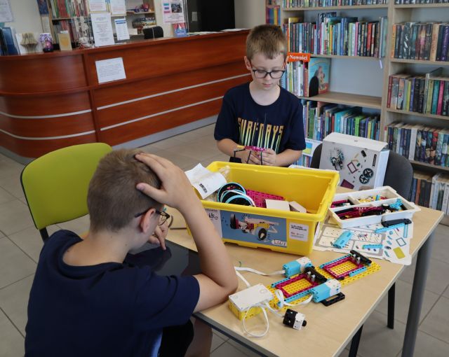 Na zdjęciu znajduje się dwóch chłopców przy stoliku, na którym znajdują się klocki LEGO, jeden z chłopców ma przed sobą tablet i koduja swojego robota. W tle znajdują się półki z książkami oraz biurko.