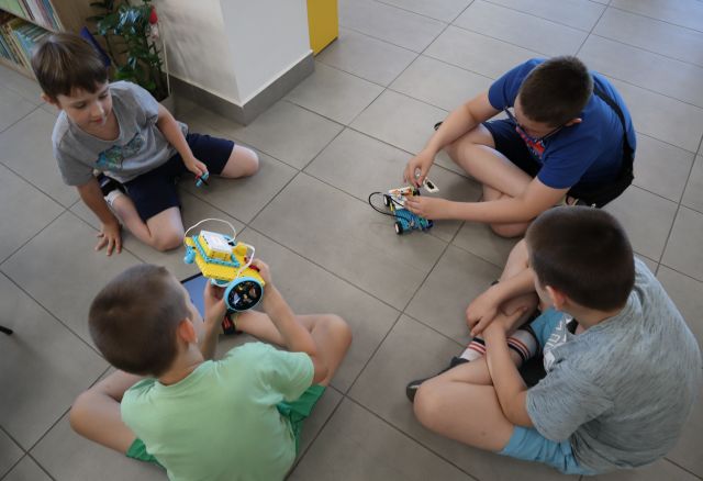 Na zdjęciu znajduje się czwórka chłopców, którzy siedzą na podłodze, jeden z nich ma na kolanach tablet, a w ręce trzyma zbudowaną z klocków LEGO konstrukcja, przed chłopcem w prawym górnym rogu znajduje się wykonany z klocków LEGO robot.
