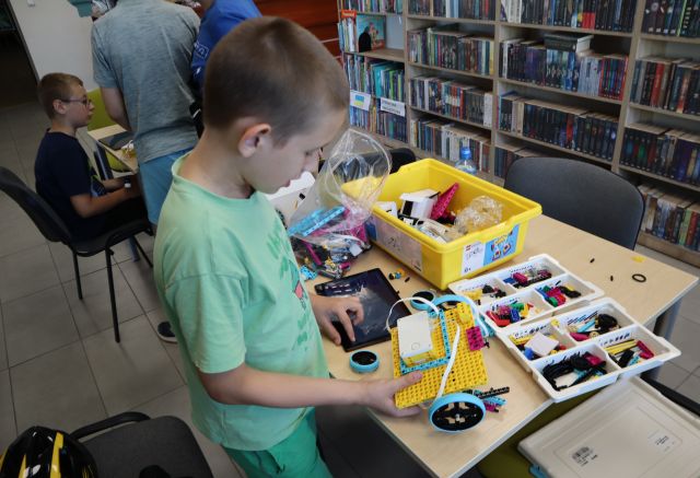 Na zdjęciu znajduje się chłopiec, przed którym na stoliku leży tablet, konstrukcja z LEGO oraz opakowanie i klocki. W tle znajduje się chłopiec przy stoliku obok oraz półki z książkami.