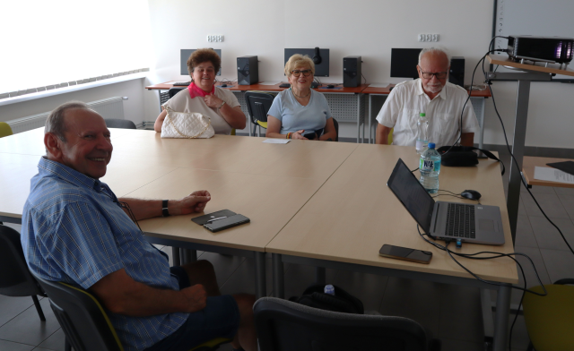 Na zdjęciu znajduje się czwórka dorosłych, którzy siedzą przy złączonych stołach w sali komputerowej, po prawej stronie leży laptop oraz jest ustawiony rzutnik.