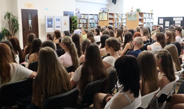 Na zdjęciu znajduje się widownia na holu biblioteki, w tle znajdują się półki z książkami, drzwi, okna.