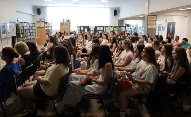 Na zdjęciu znajduje się widownia na holu biblioteki, głównie młodzież , w tle znajdują się okna, półki z książkami, tablice. Zdjęcie zostało wykonane od boku.