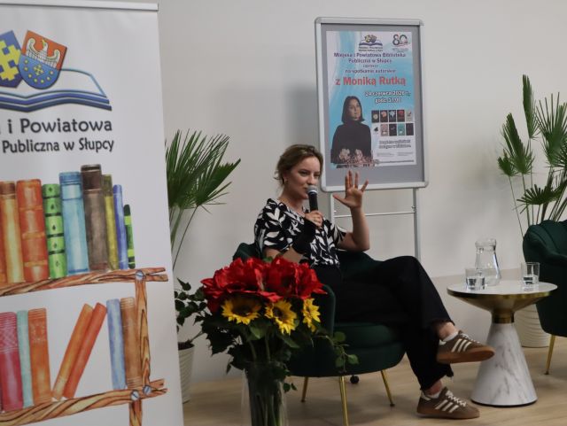 Na zdjęciu znajduje się kobieta na scenie w jednej ręce trzyma mikrofon, drugą ma podniesioną do góry. Za nią znajdują się rośliny oraz plakat promujący spotkanie, po lewej stronie znajduje się baner z logiem biblioteki i książkami.
