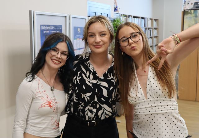 Na zdjęciu znajdują się dwie dziewczyny stojące do zdjęcia wraz z autorką. W tle znajdują się plakaty oraz półki z książkami.