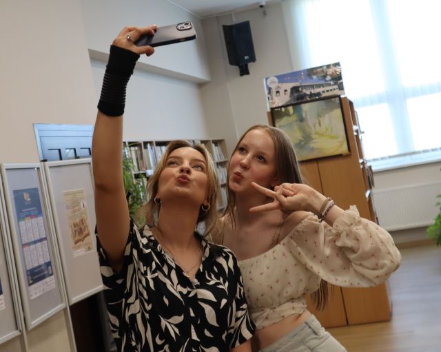 Na zdjęciu znajduję się kobieta trzymająca telefon do góry i robiąca zdjęcie ze stojącą koło niej nastolatką. W tle znajdują się plakaty i półki z książkami.