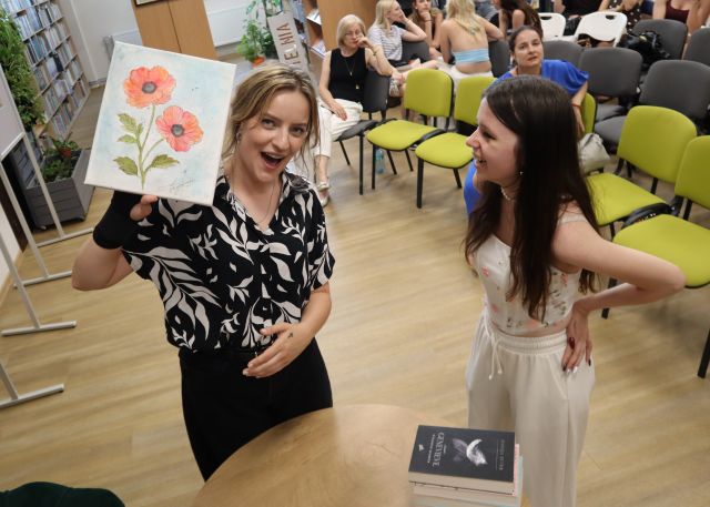 Na zdjęciu znajduję się dziewczyna po prawej i kobieta po lewej, która pokazuje do kamery obraz wykonany przez nastolatkę. W tle znajdują się krzesła i inni uczestnicy. 