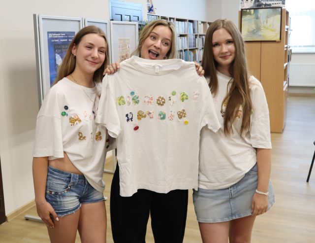 Na zdjęciu znajdują się dwie dziewczyny w takich samych koszulkach, na środku znajduje się kobieta, która trzyma przed sobą taką samą koszulkę. W tle znajdują się półki z książkami i plakaty.