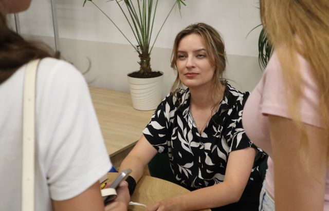 Na zdjęciu znajduje się kobieta przy stoliku, a przed nią stoją dwie dziewczyny. W tle znajduje się scena, na której stoi kwiat.