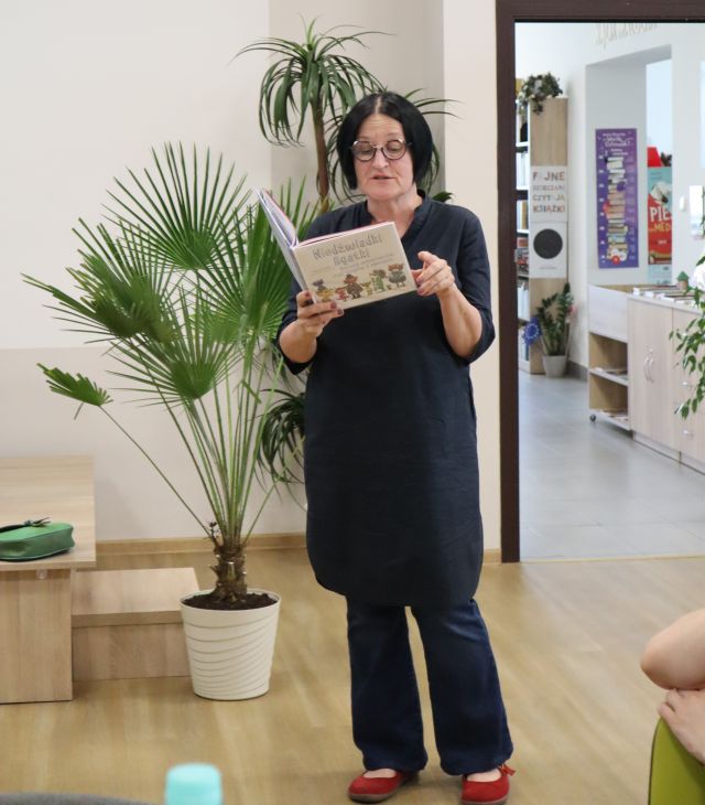 Na zdjęciu znajduje się kobieta czytająca książkę. W tle znajdują się kiwaty, drzwi do sali bibliotecznej. 