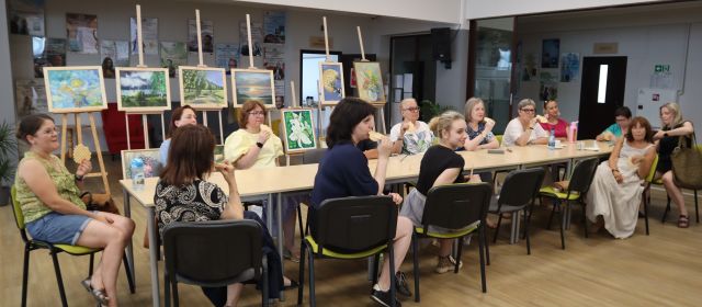 Na zdjęciu znajdują się kobiety siedzące przy złączonych stołach na holu biblioteki. Kobiety jedzą wafelki. W tle znajdują się obrazy.