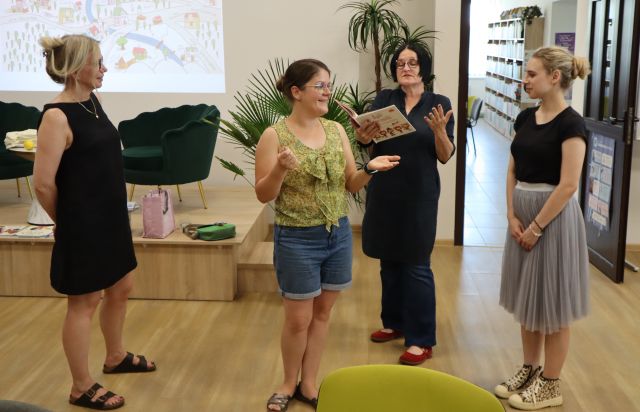 Na zdjęciu znajdują się cztery kobiety, jedna z nich czyta książkę. W tle znajduje się scena z fotelami, kawałek wyświetlanej na ścianie prezentacji oraz drzwi do sali bibliotecznej po prawej stronie.